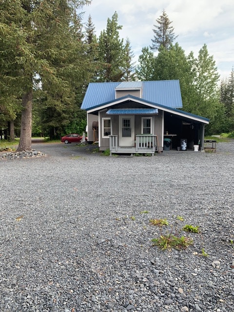 A two-story house with a blue metal roof is positioned on gravel space. A porch with a railing frames the entrance, surrounded by trees. A vehicle is parked nearby, with a yard featuring patches of grass and rocks.