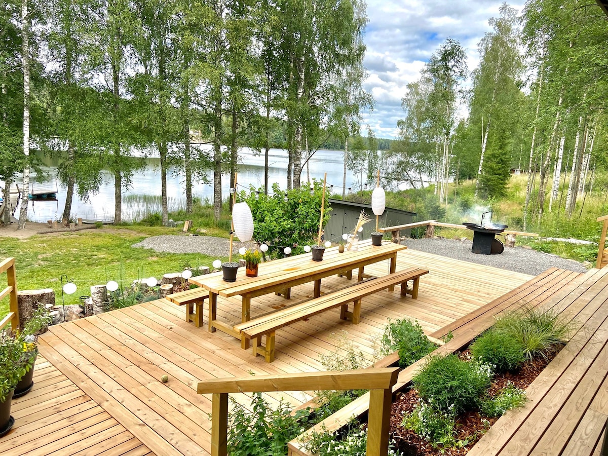 A spacious wooden deck is showcased with long seating and a dining table, surrounded by lush greenery and potted plants. A view of the serene lake and forest can be seen in the background, while a fire pit is positioned nearby for gatherings.