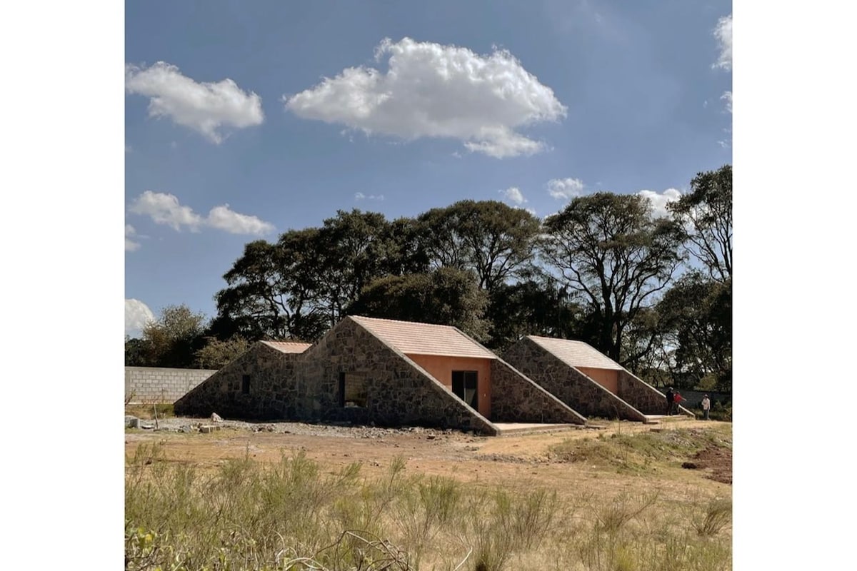 A series of modern, angular stone structures are set against a clear blue sky. The buildings exhibit earthy tones, blending with the natural surroundings. Lush trees provide shade, creating a tranquil environment, while the open landscape hints at ample space for outdoor activities.