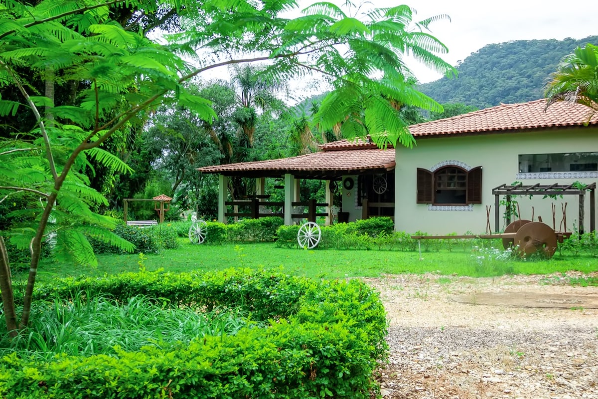 A tranquil landscape is presented, featuring a charming home with a tiled roof surrounded by lush greenery. A well-maintained garden bed, shaped like a circle, highlights the vibrant plants and trees, with a backdrop of distant mountains providing a serene setting.