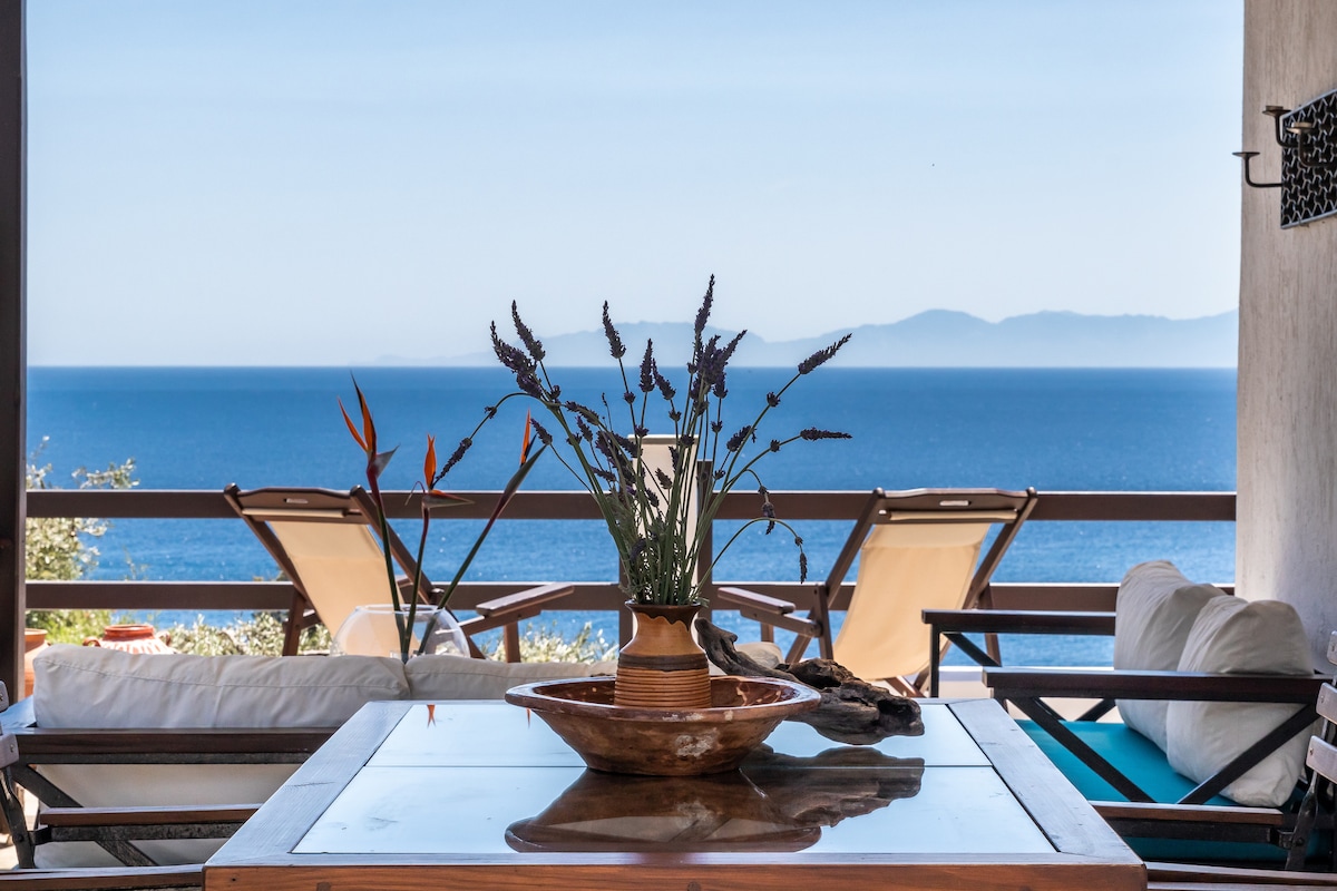 A terrace space features a wooden table with a decorative bowl and a vase holding lavender. Two lounge chairs are positioned nearby, overlooking the expansive sea and distant mountains. A sofa with soft cushions offers additional seating, enhancing the relaxing atmosphere.