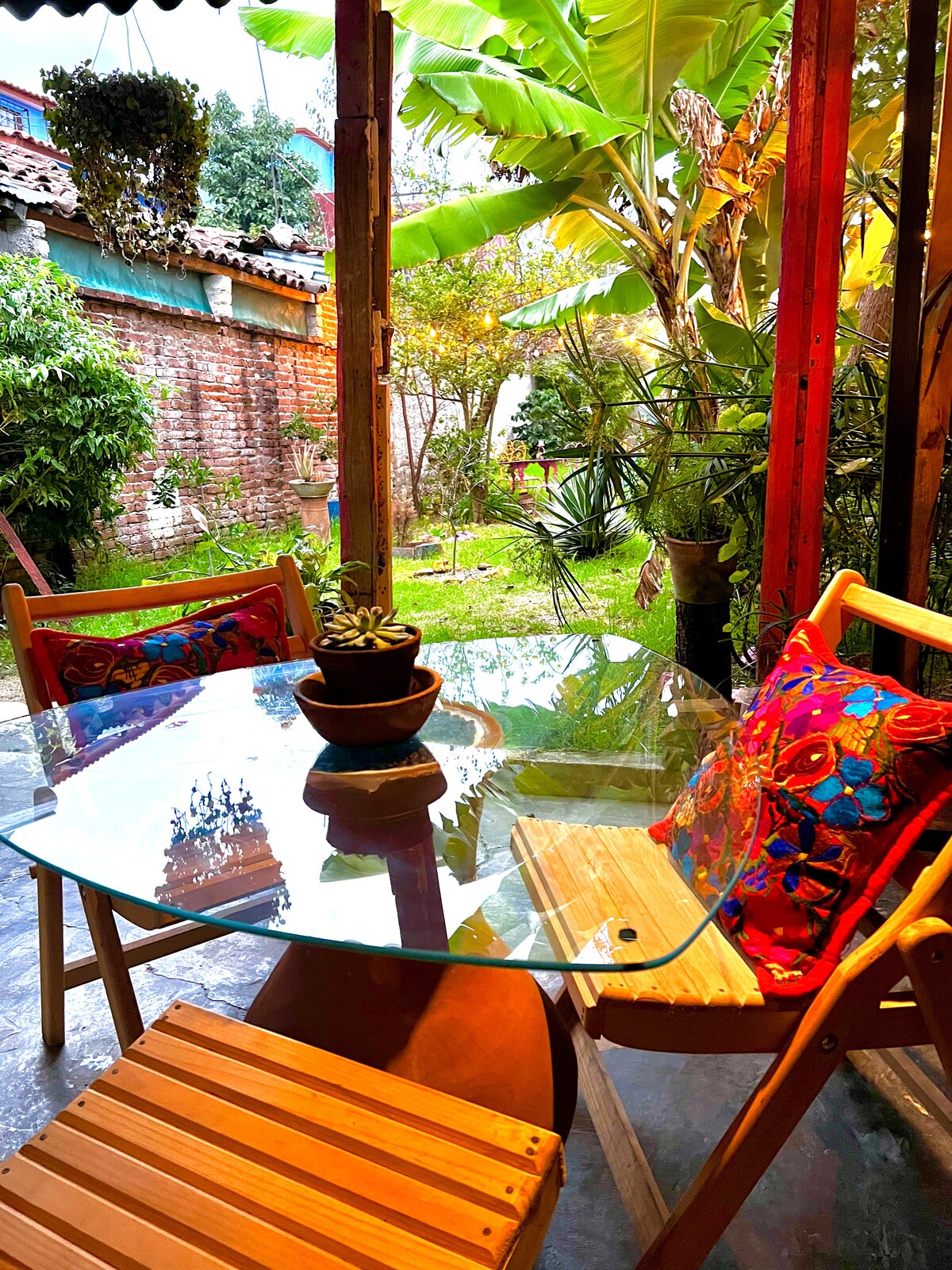 A clear glass table is set in an outdoor area, accompanied by two wooden chairs adorned with colorful cushions. Lush greenery and palm plants surround the space, with a small garden visible in the background, creating a serene atmosphere for relaxation.