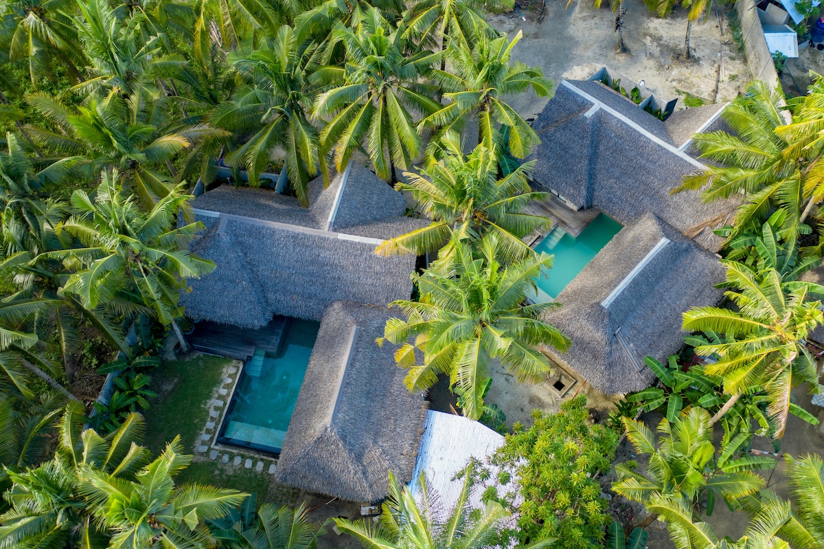 An aerial view captures a private villa surrounded by lush palm trees. The structures feature traditional thatched roofs, and a private swimming pool is visible between them. Natural elements blend harmoniously with the landscape, creating a serene and tropical environment.