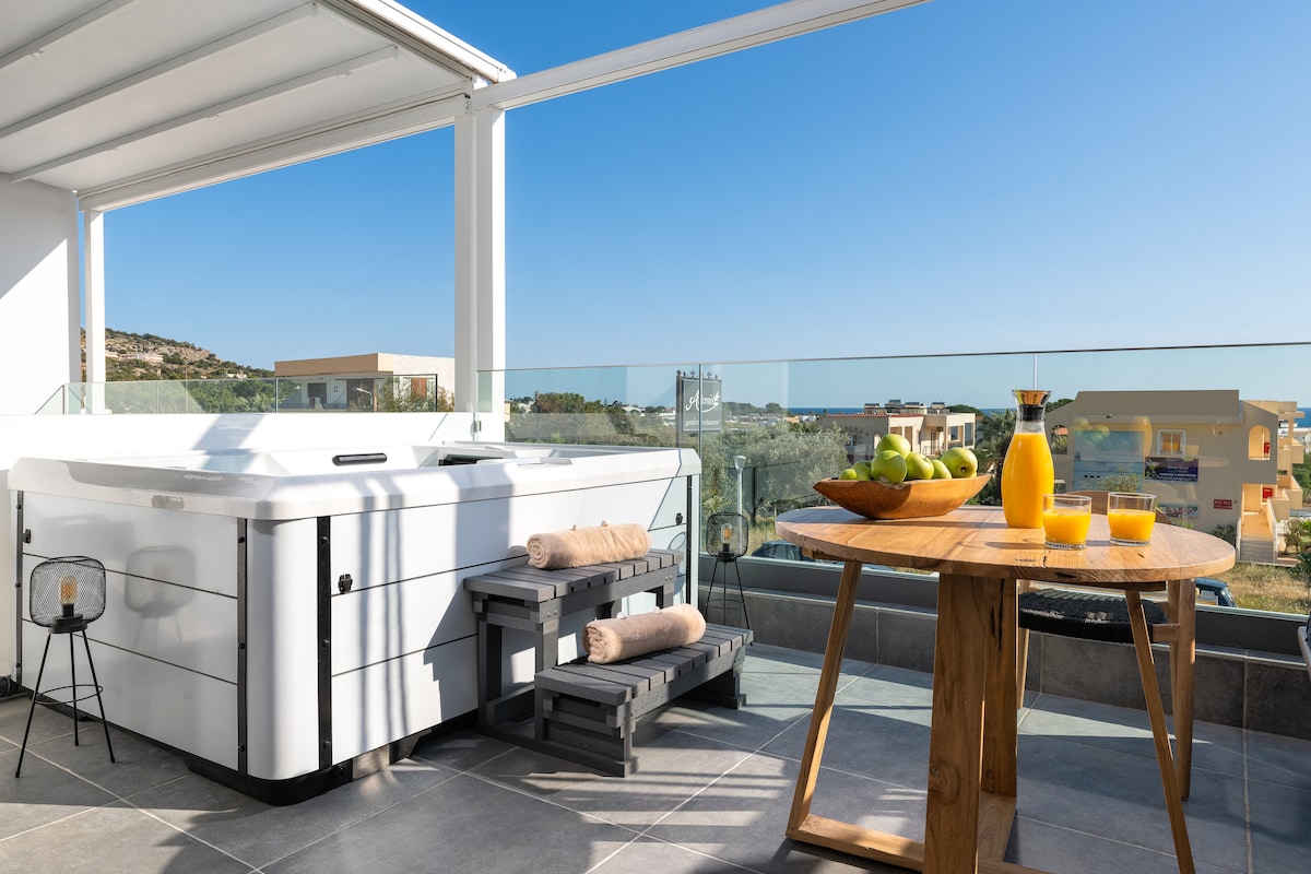 An outdoor private veranda showcases a modern hot tub, complemented by wooden steps and a small side table. A round wooden dining table is set with a fruit bowl and drinks, surrounded by clear views of the landscape under a bright blue sky.