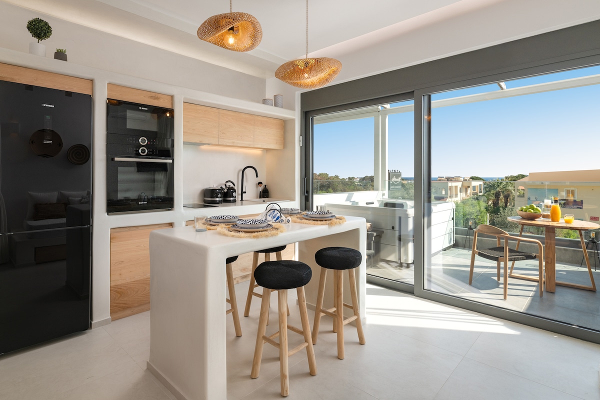 A modern kitchen space features a sleek black refrigerator and built-in oven, paired with wooden cabinetry. A central island with stools provides seating, while large glass sliding doors open to a balcony with outdoor dining, offering views of the surrounding area.