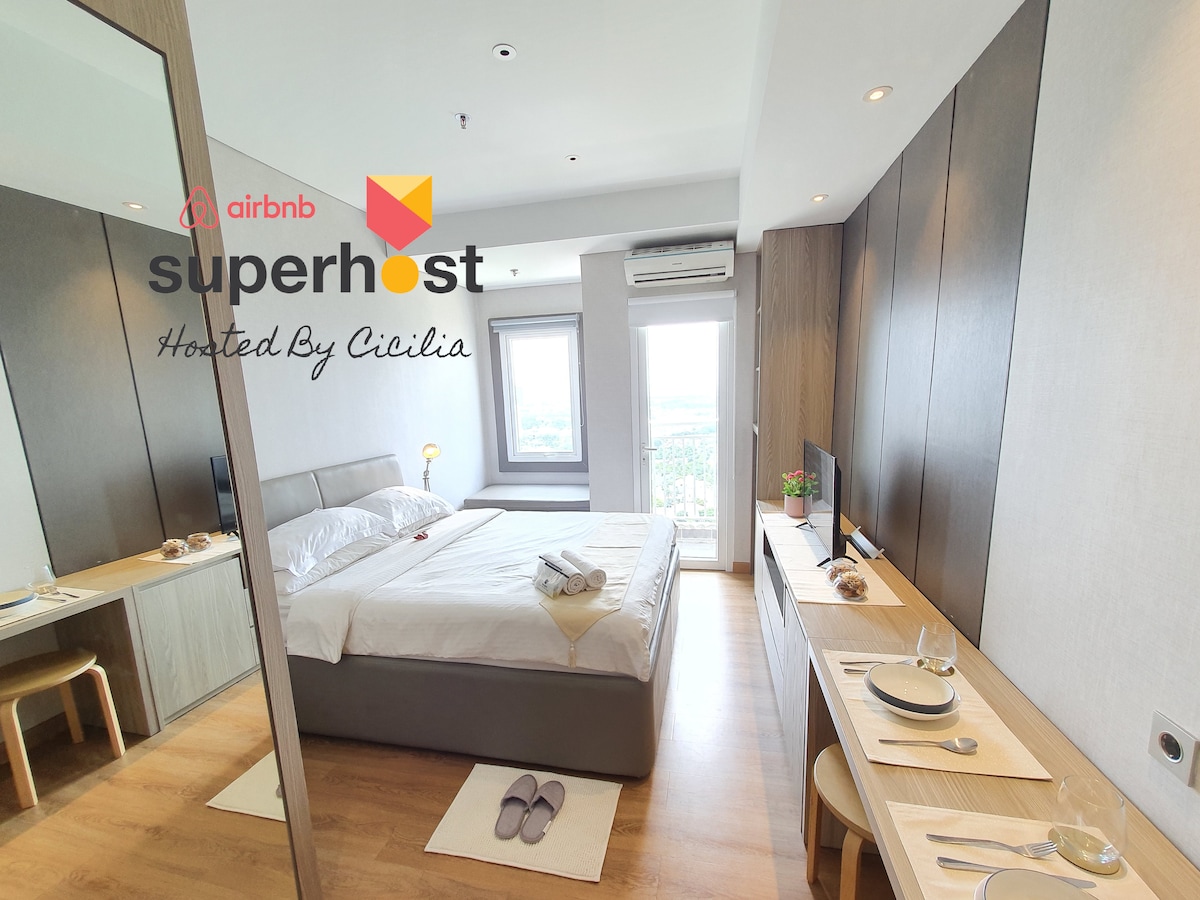 A modern studio apartment is shown, featuring a spacious bed topped with white linens and a light blanket. A dressing table and mirror reflect the tidy, neutral-toned decor. Natural light fills the room through a window, offering a view of the outside. Dining utensils are neatly arranged on the table.