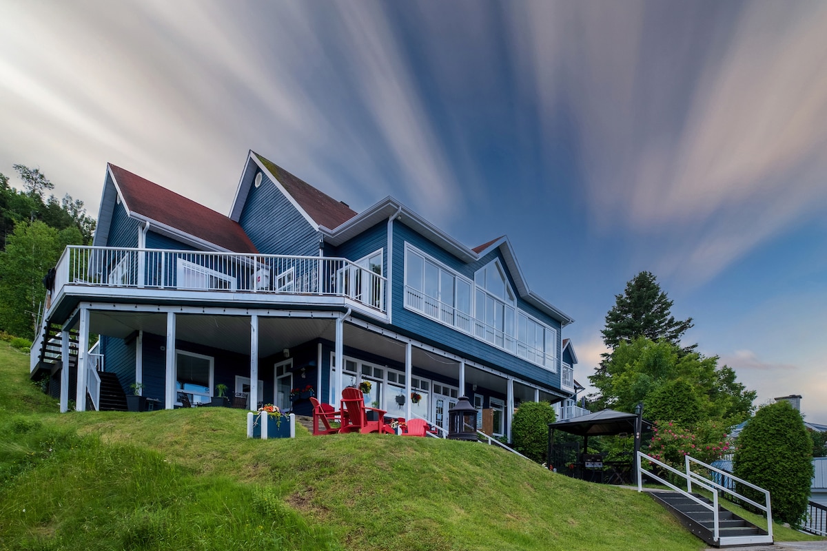 The exterior of the condo features a spacious deck with seating arrangements, including red chairs. A grassy area surrounds the building, enhancing its inviting appearance. The structure showcases large windows and a triangular roof, set against a backdrop of a dynamic sky.