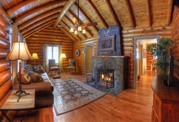 A spacious living area features vaulted log ceilings and a stone fireplace that provides a warm ambiance. Comfortable seating options are arranged around a patterned area rug. Windows allow natural light to fill the space, and a doorway leads to additional areas of the cabin.