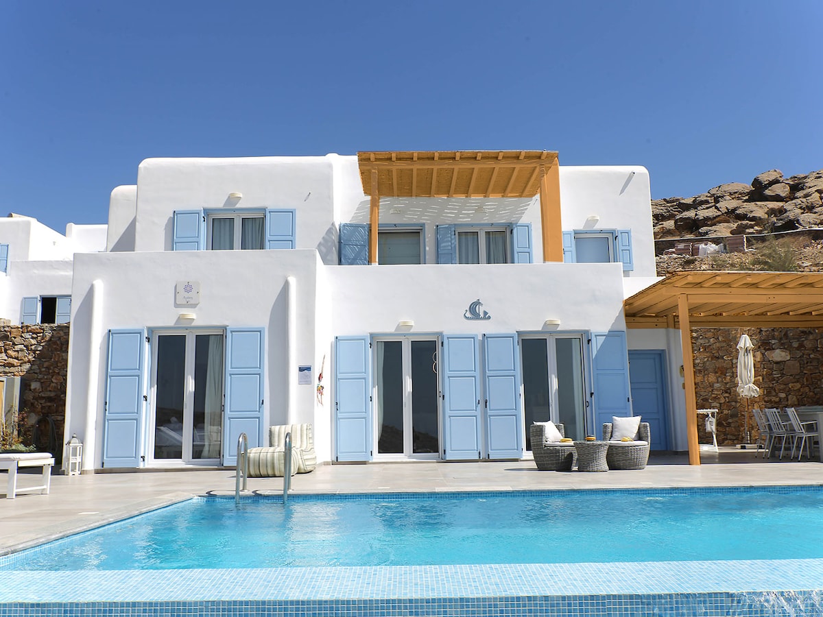 The exterior of the villa displays traditional Cycladic architecture, featuring light blue shutters and a warm wooden pergola. The private pool is visible in the foreground, accompanied by stylish sun loungers. Clear skies enhance the vibrant ambiance, offering views of the surrounding hillside.