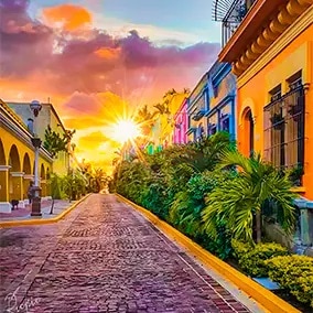 A vibrant street is captured at sunset, with colorful buildings flanking the cobblestone pathway. Lush tropical plants add greenery along the sides. The sun's rays create a warm glow, enhancing the inviting scene of the picturesque neighborhood.