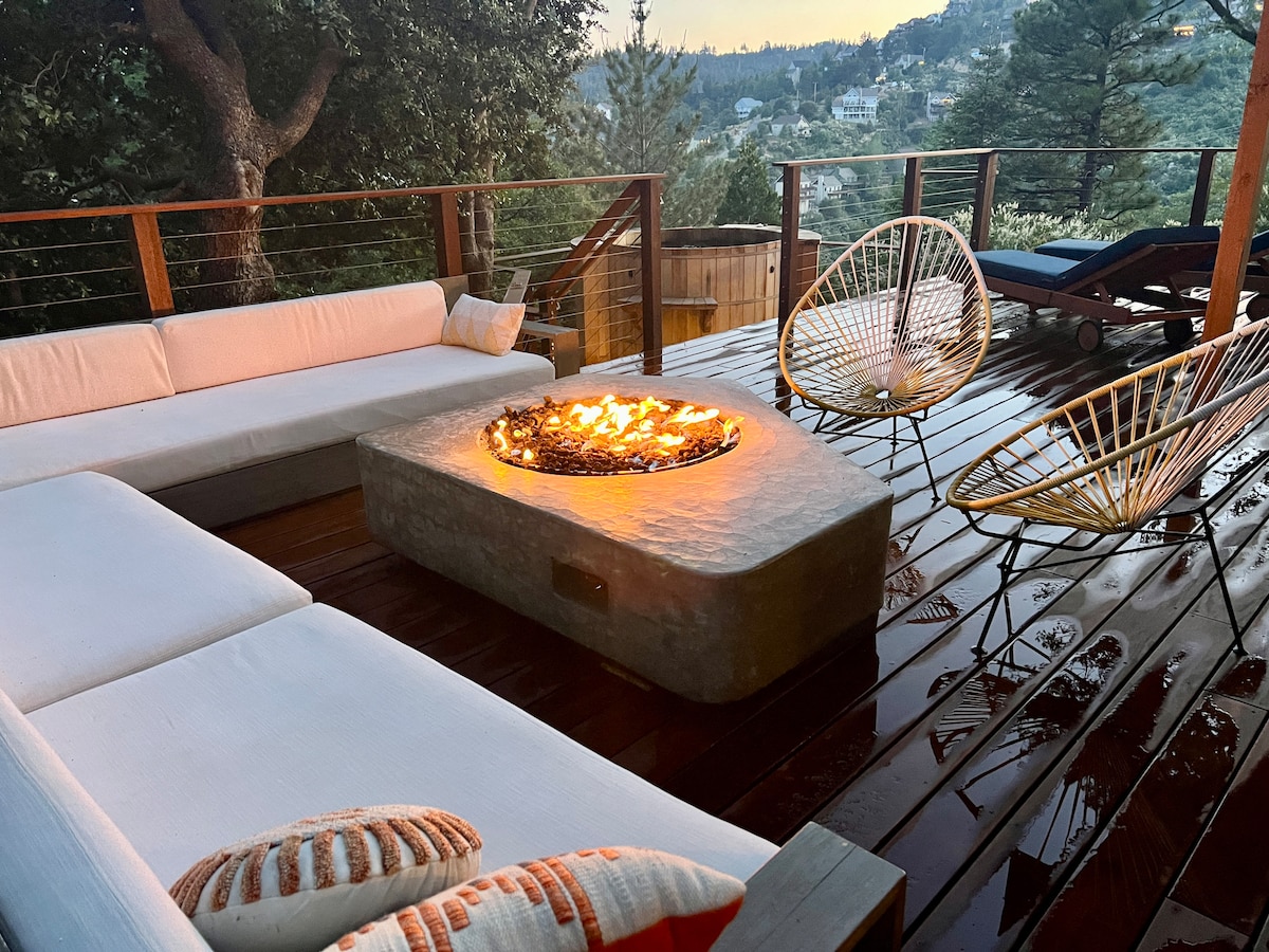 An outdoor deck is furnished with a modern concrete gas fire pit at the center, surrounded by stylish seating. The area features a mix of comfortable couches and accent chairs, providing a cozy space for gatherings. Wood decking and views of the surrounding trees are visible.