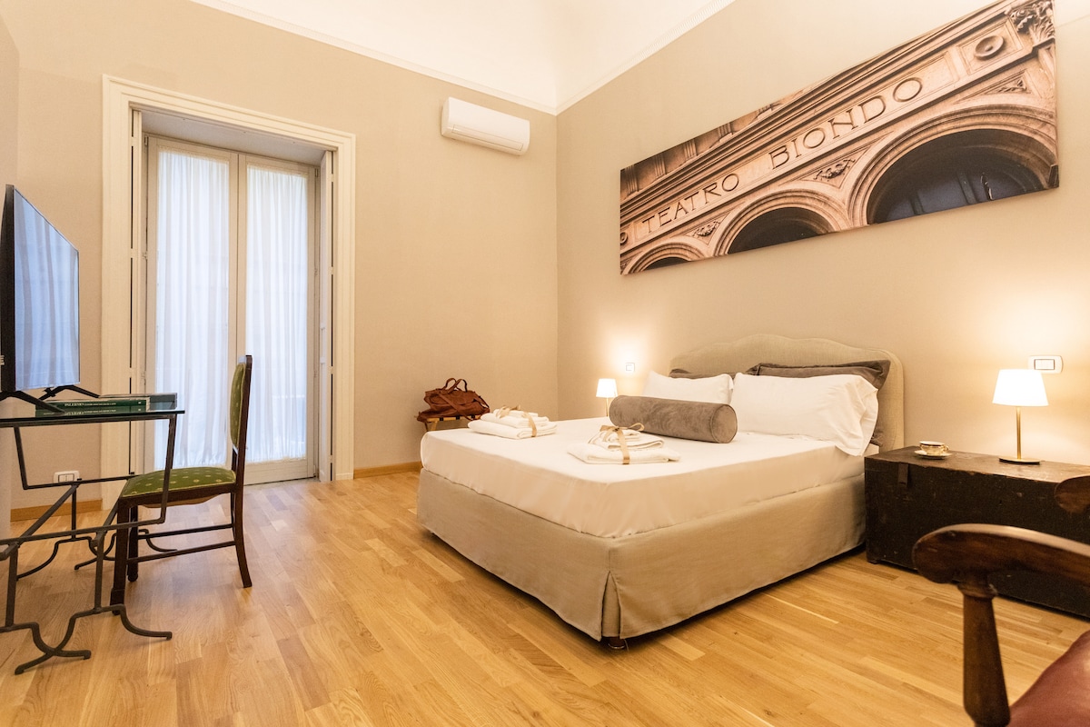 A spacious room features a double bed with neutral bedding, a wooden desk with a chair, and a television on an adjacent stand. Large windows are dressed with sheer curtains, enhancing the airy vibe. A decorative wall art piece of the Teatro Biondo adds an elegant touch.