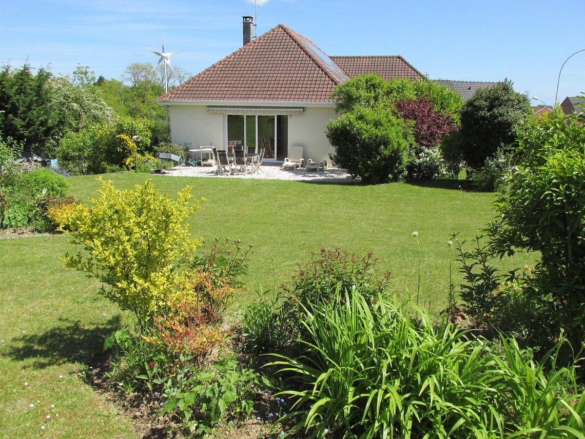The image features a single-story house surrounded by a well-maintained garden. The green lawn is complemented by varied shrubs and flowers. A patio area, equipped with seating, is visible at the back of the house, offering a peaceful outdoor space.