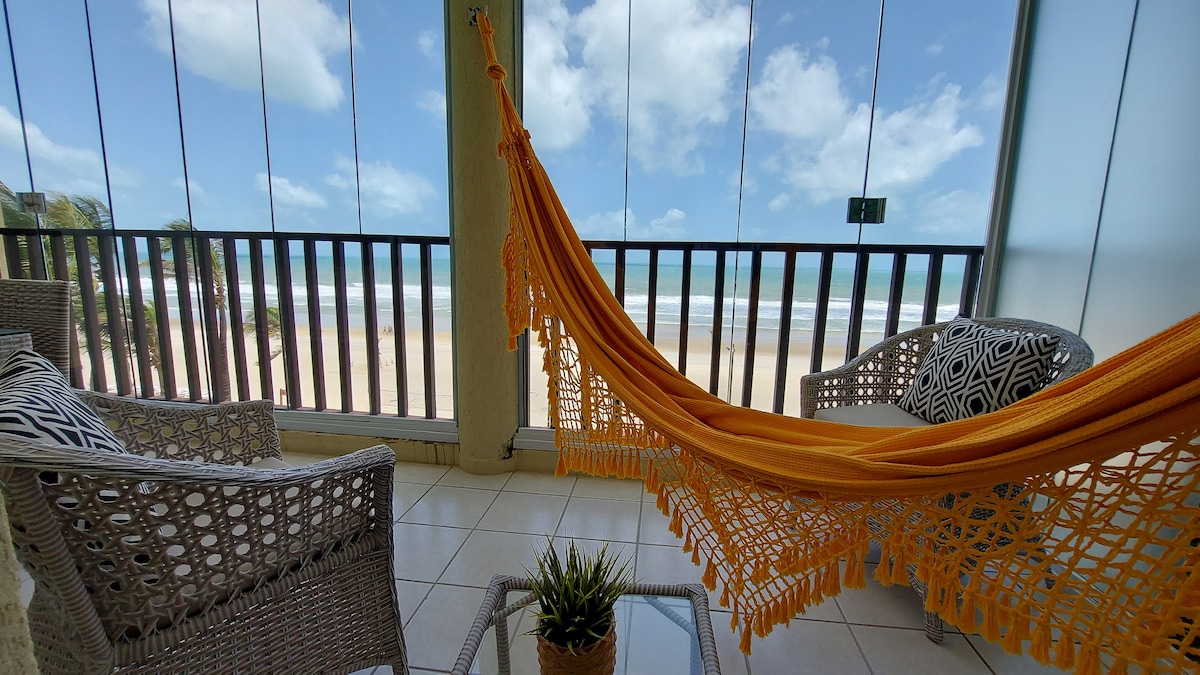 A balcony features a vibrant orange hammock, inviting relaxation. Two woven chairs are positioned beside a small glass table with a potted plant. Floor-to-ceiling windows provide panoramic views of the beach and ocean, allowing abundant natural light into the space.