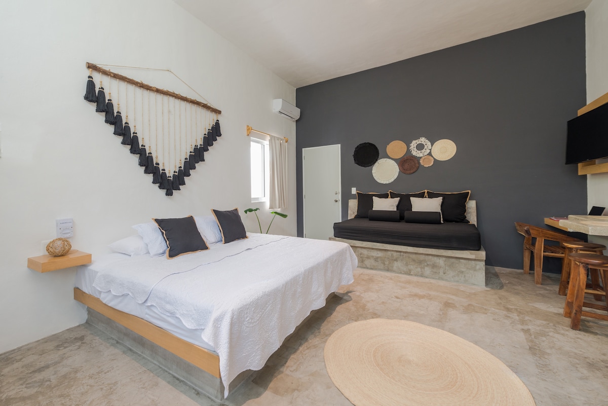A spacious room features a king-sized bed with crisp linens and dark accent pillows. A separate seating area includes a black sofa and decorative wall hangings. Natural light enters through the window, illuminating a round rug placed on a polished concrete floor.