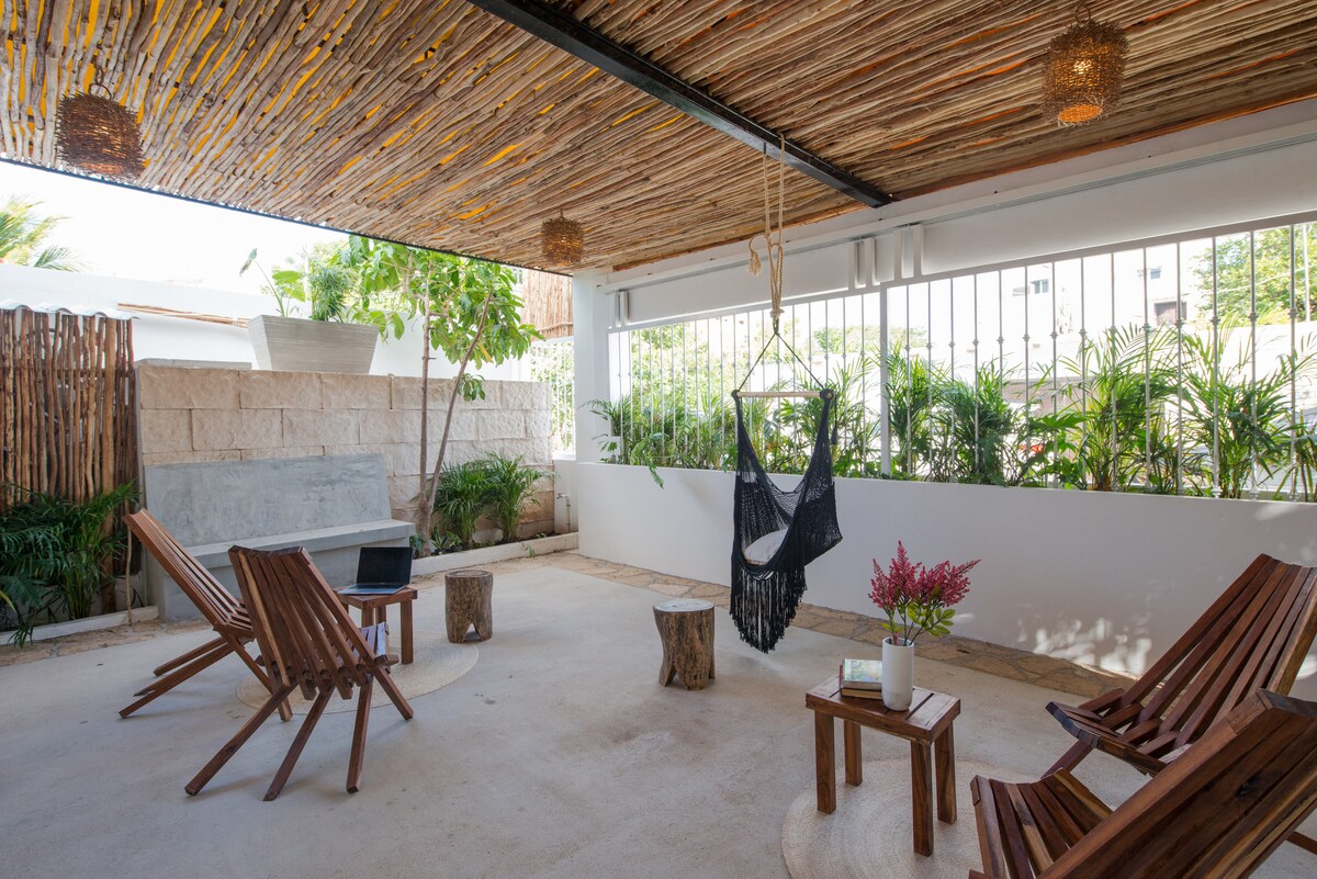An outdoor shared terrace is shown with a natural design, featuring wooden lounge chairs and a small coffee table. A hanging chair is suspended nearby, while greenery adds a refreshing touch to the space. Soft lighting creates a cozy atmosphere beneath the textured ceiling.