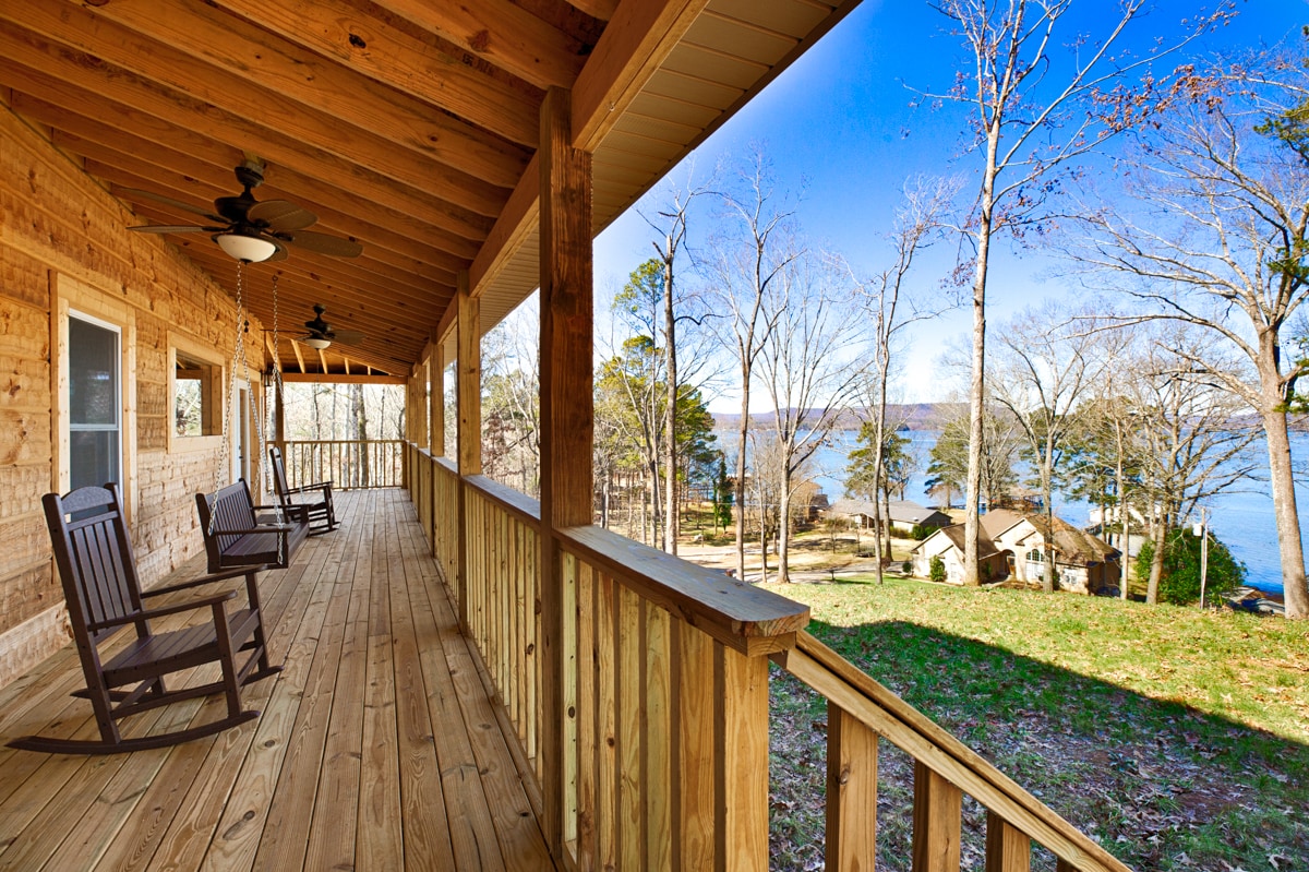 A serene wrap-around porch features wooden rocking chairs, providing a relaxing space to enjoy views of the surrounding landscape and the nearby lake. Bare trees reflect the tranquil setting, while distant houses hint at the community. The expansive deck showcases natural wood tones.