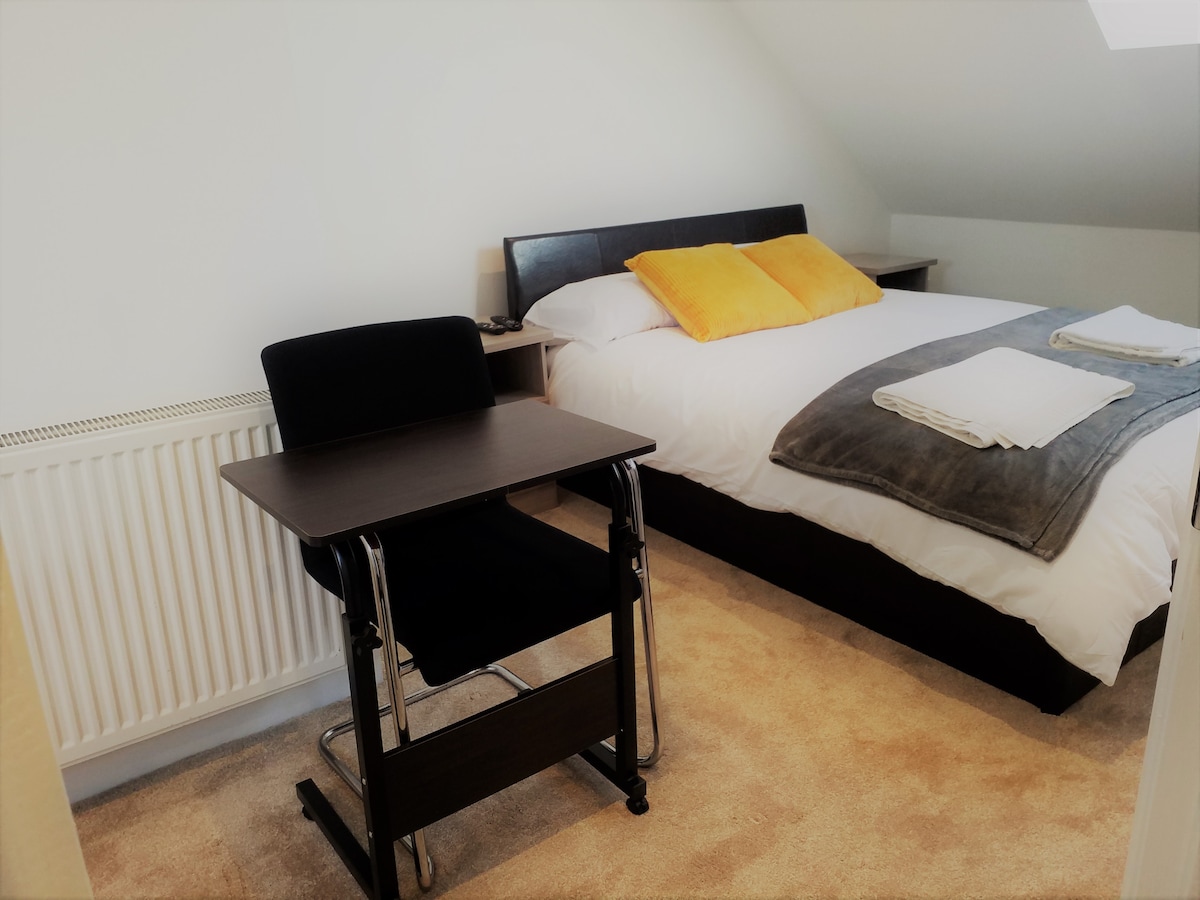 A well-lit sleeping area features a spacious bed with neutral linens and two yellow accent pillows. A modern desk with a chair is positioned nearby, alongside a radiator. Soft carpet covers the floor, contributing to a cozy atmosphere.