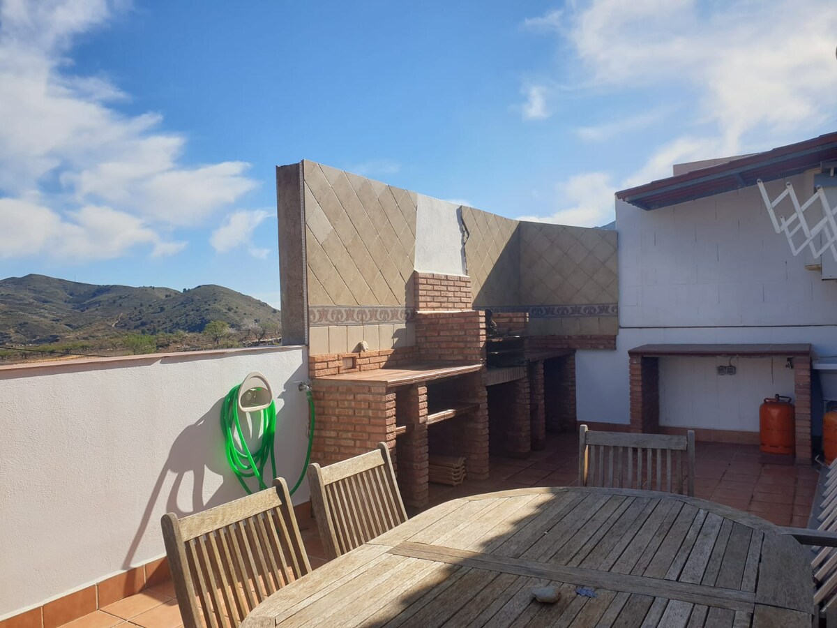 An outdoor terrace features a rustic wooden table surrounded by several wooden chairs. A built-in BBQ area with brick detailing is visible along one side, complemented by views of rolling hills under a clear blue sky.