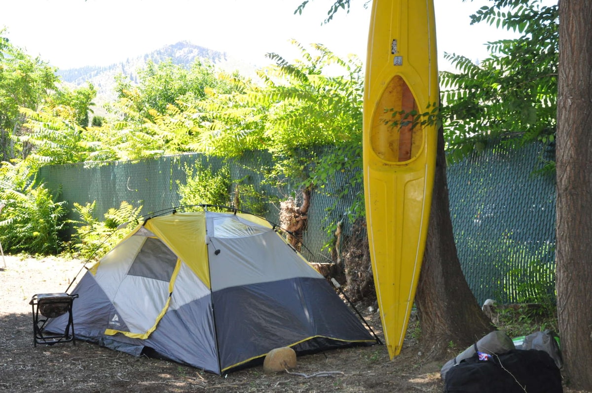 A spacious tent is set up on a gravel surface, surrounded by lush greenery and a protective fence. A bright yellow kayak hangs from a tree, adding a pop of color to the scene. Camping gear is neatly placed nearby, indicating a cozy and functional camping area.