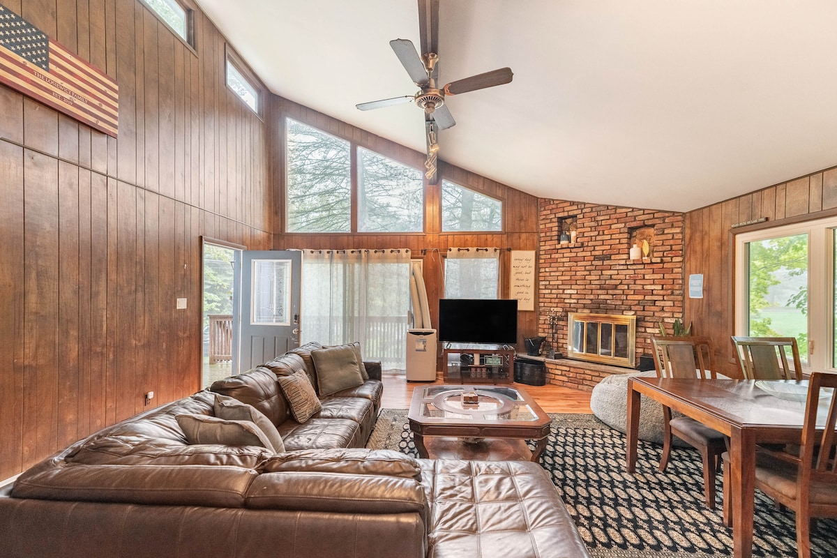 A spacious living area features a large brown leather sectional sofa and a wooden dining table. A brick fireplace serves as a focal point, while large windows allow natural light to fill the room. A television is positioned near the seating, contributing to a cozy atmosphere.