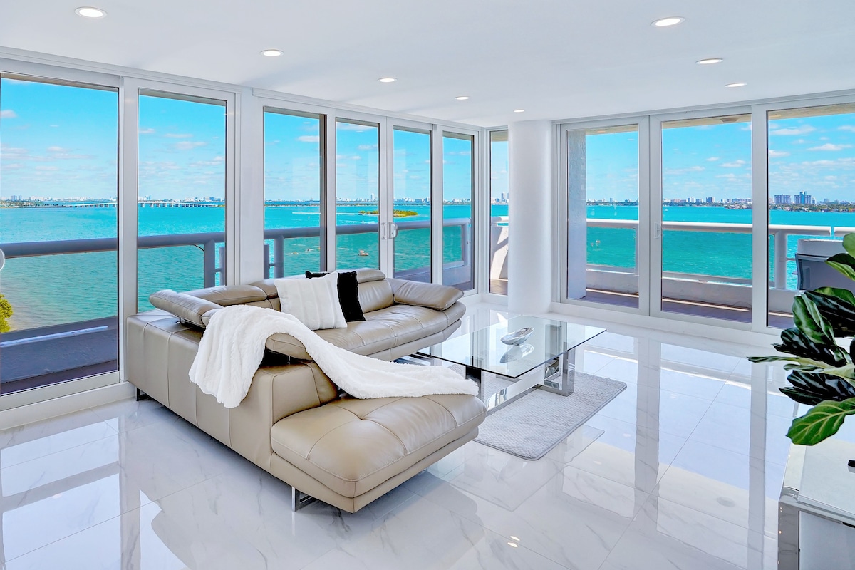 A spacious living area features a light-toned leather sectional sofa, positioned to face large floor-to-ceiling windows. The room allows for abundant natural light, showcasing expansive views of Biscayne Bay and distant Miami skyline. A sleek glass coffee table sits prominently in front.