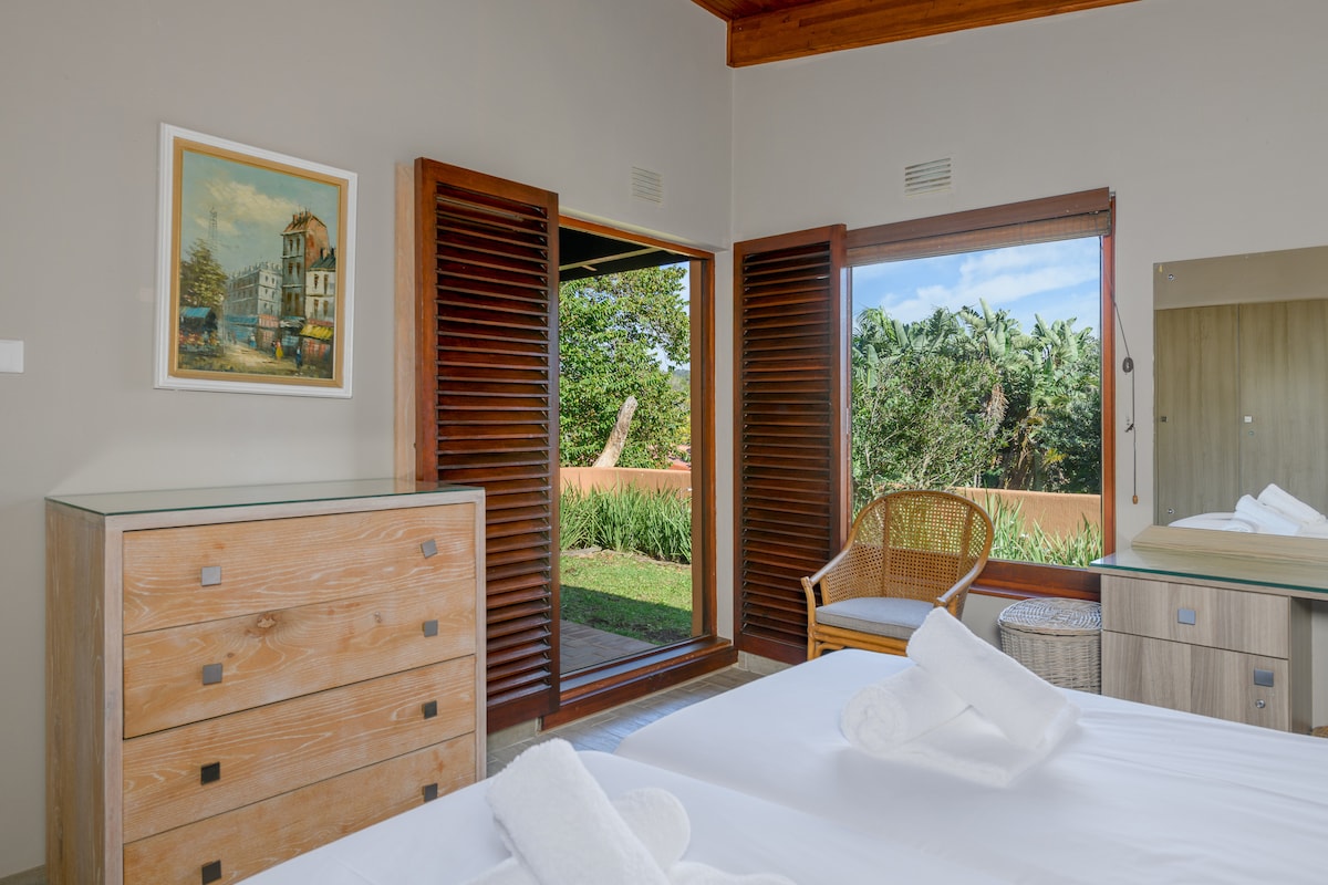 A bright bedroom features two single beds with neatly arranged white towels. A wooden dresser is positioned beside a chair, while large windows with wooden shutters offer a view of the lush greenery outside.