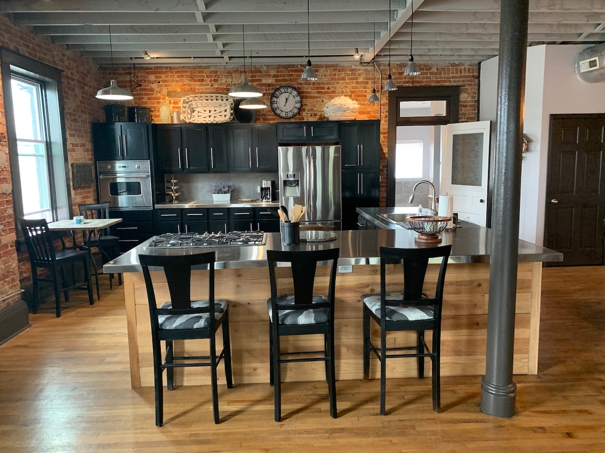 A modern kitchen features a spacious island with three black chairs. The stainless steel appliances are complemented by dark cabinetry and exposed brick walls, providing a rustic yet contemporary feel. Large windows allow natural light to brighten the area, enhancing the inviting atmosphere.