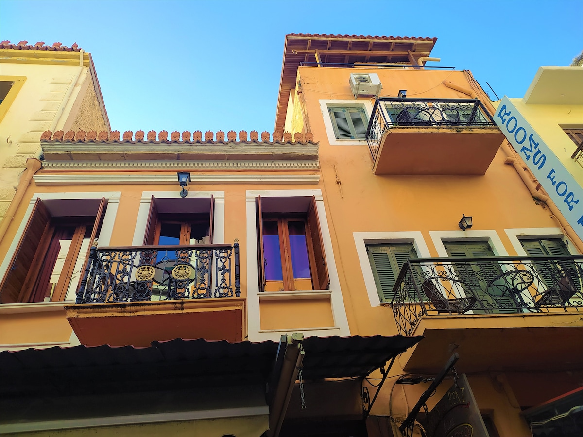An exterior view of a charming building features traditional balconies adorned with ornate iron railings. Windows are framed with wooden shutters, reflecting the vibrant colors of Chania Old Town, while a clear blue sky adds to the inviting atmosphere.