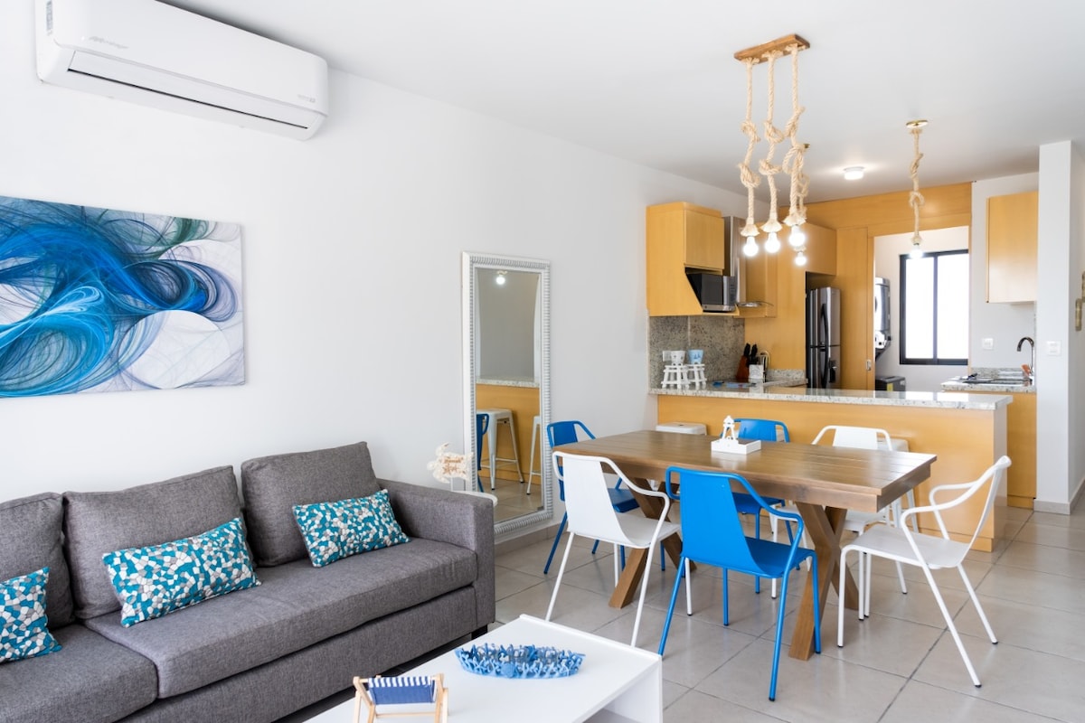 A modern living space is shown, featuring a cozy gray sofa with patterned throw pillows. A dining table with white and blue chairs is positioned adjacent to a stylish kitchen with wooden cabinets, while a large mirror and artwork add visual interest to the room.