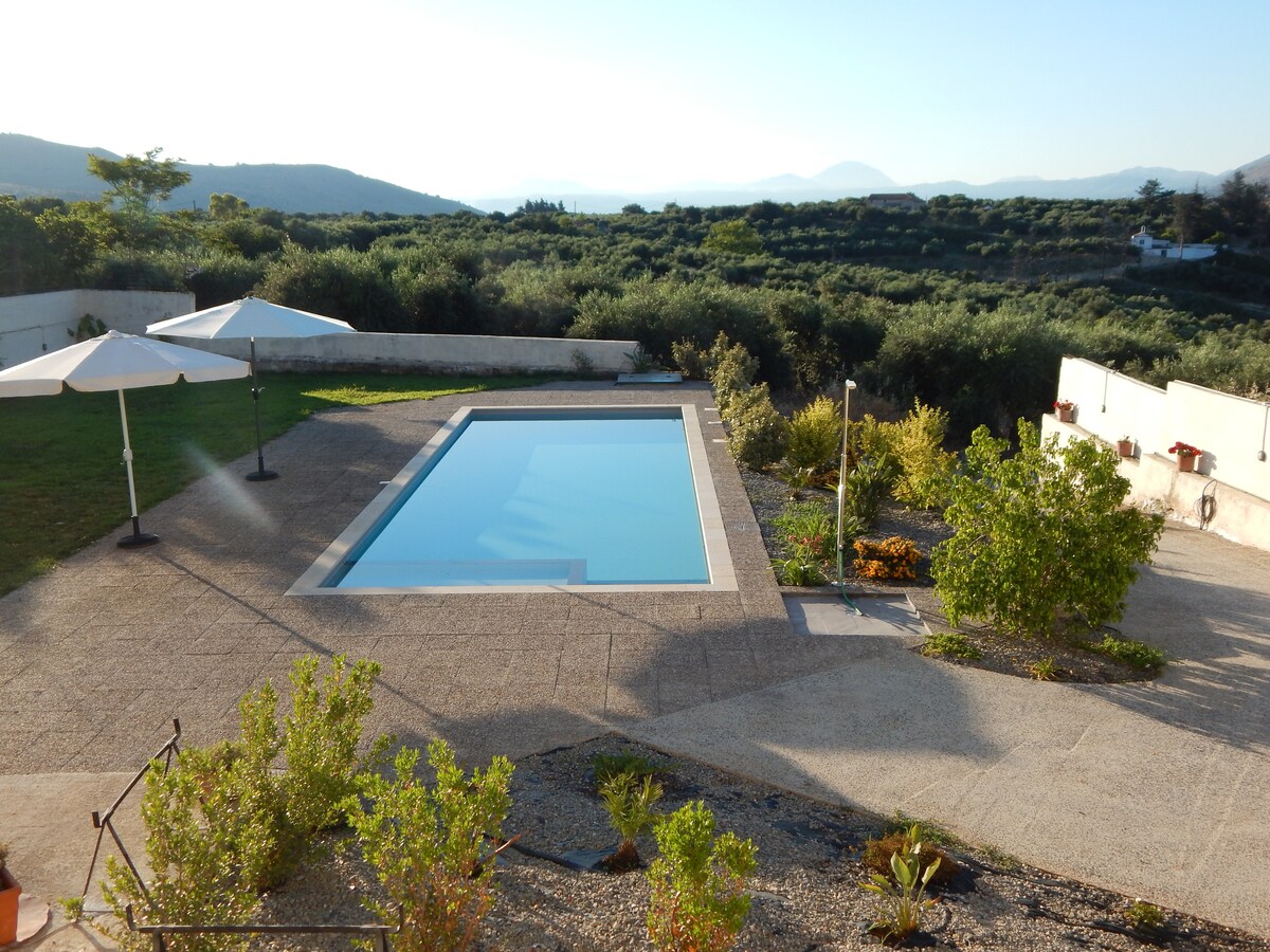 A clear blue pool is situated within a sunlit outdoor area, surrounded by well-maintained gardens. Two umbrellas provide shade beside the pool, while distant hills frame the serene landscape.