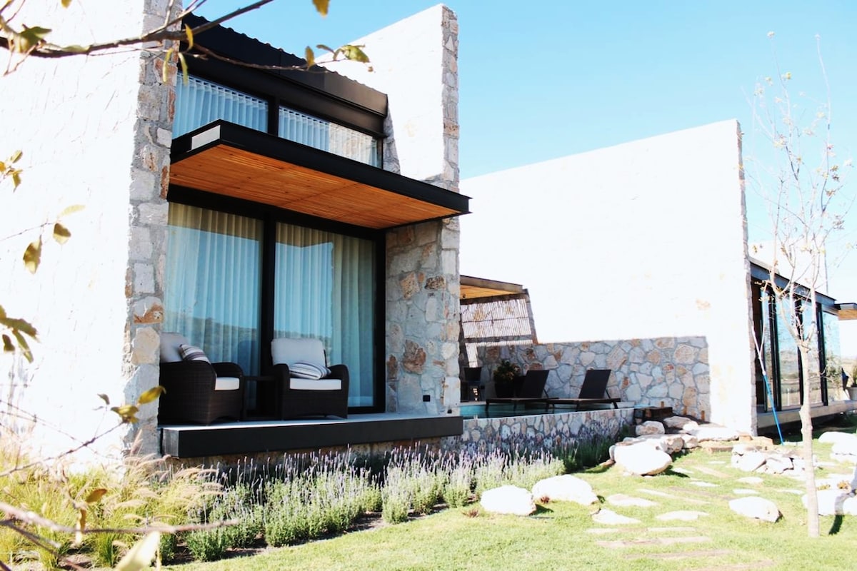 A modern villa features natural stone exteriors and large glass windows. The outdoor area includes a cozy seating arrangement with two chairs on a wooden deck. Lush greenery and decorative stones outline the space, creating a serene environment.