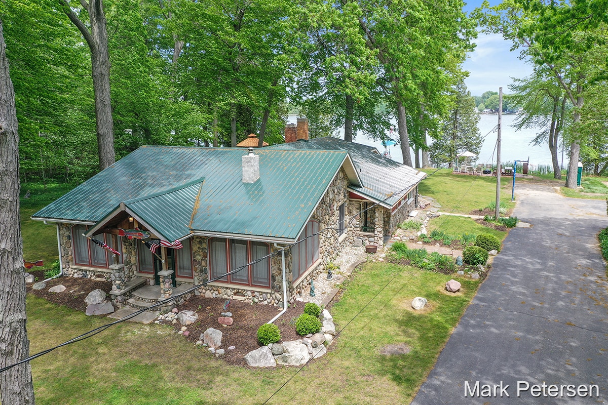 The Stony Lodge is set among tall trees, featuring a stone exterior and a sloped metal roof. A well-maintained lawn and a paved driveway lead to the entrance. The background reveals Clifford Lake's serene waters, enhancing the setting's natural beauty.