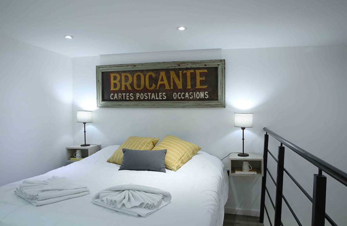 A well-lit bedroom features a spacious bed with soft linens and decorative pillows. Two bedside lamps provide gentle lighting. A vintage-style sign is mounted above the bed, contributing to the room's character.