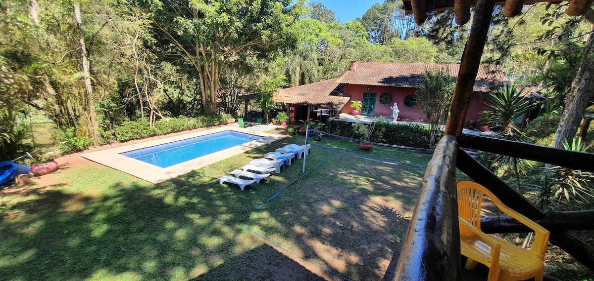 An aerial view of the outdoor area showcases a spacious swimming pool surrounded by green grass and lounge chairs. The rustic house and trees create a natural setting, with a pathway visible leading towards the entrance. Bright yellow chairs are placed nearby for relaxation.