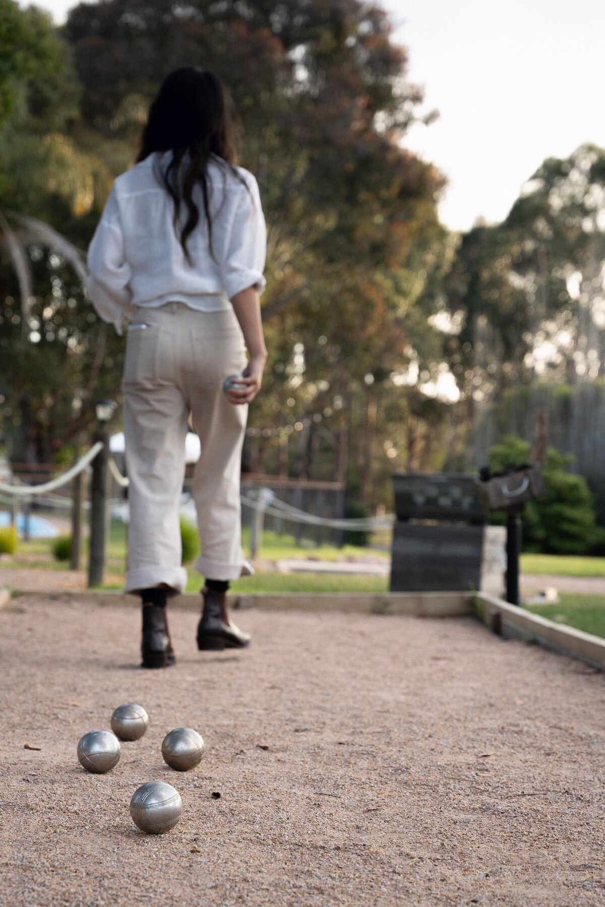 A guest is seen playing with metallic pétanque balls on a gravel surface, with trees and greenery in the background. The guest is dressed in a light-colored outfit, walking along the path, engaging in this outdoor activity that promotes leisure and social interaction.