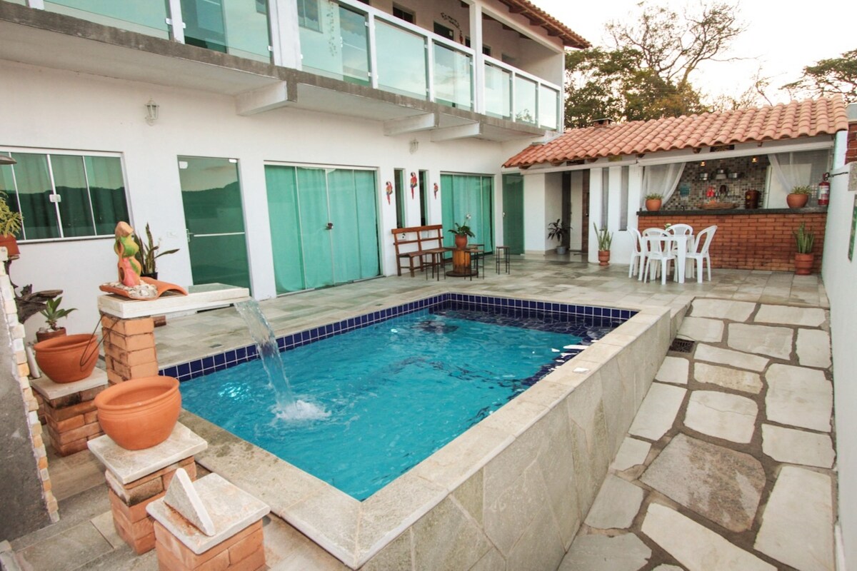 An inviting outdoor area features a blue swimming pool with a waterfall, surrounded by stone tiles and potted plants. A dining set with four chairs is positioned nearby, while additional seating is provided by a wooden bench. A covered space can be seen in the background.