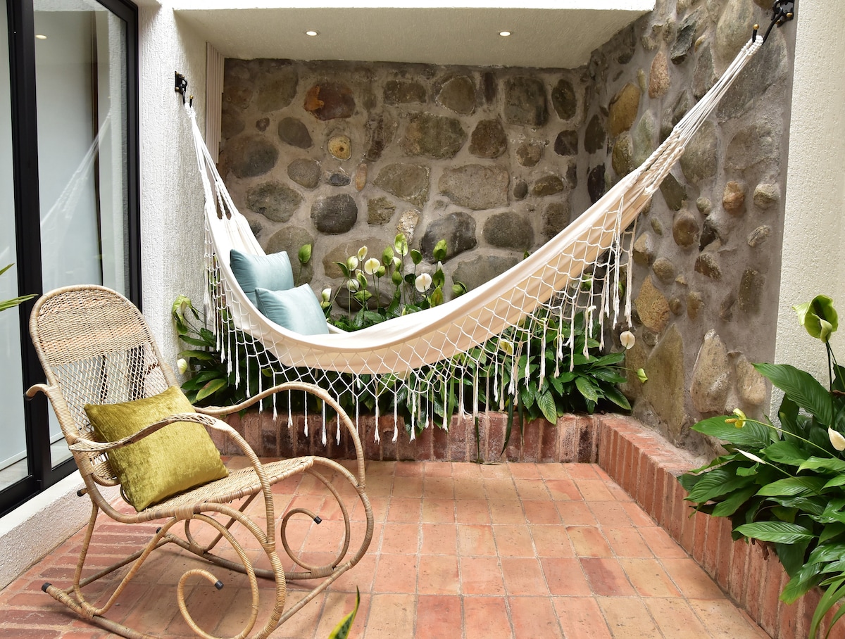 A serene private terrace features a hanging hammock against a backdrop of stone walls. Lush green plants surround the space, while a vintage rocking chair with a soft green cushion invites relaxation. Natural light brightens the area, creating a peaceful atmosphere.