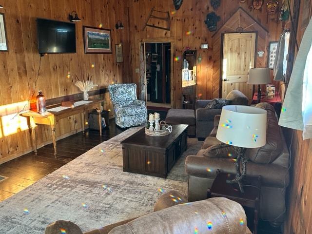 A spacious great room features wooden walls and floors, creating a warm environment. Comfortable seating is arranged around a central coffee table. A television is mounted on one wall, while natural light filters through nearby windows, illuminating the rustic decor and accessories.