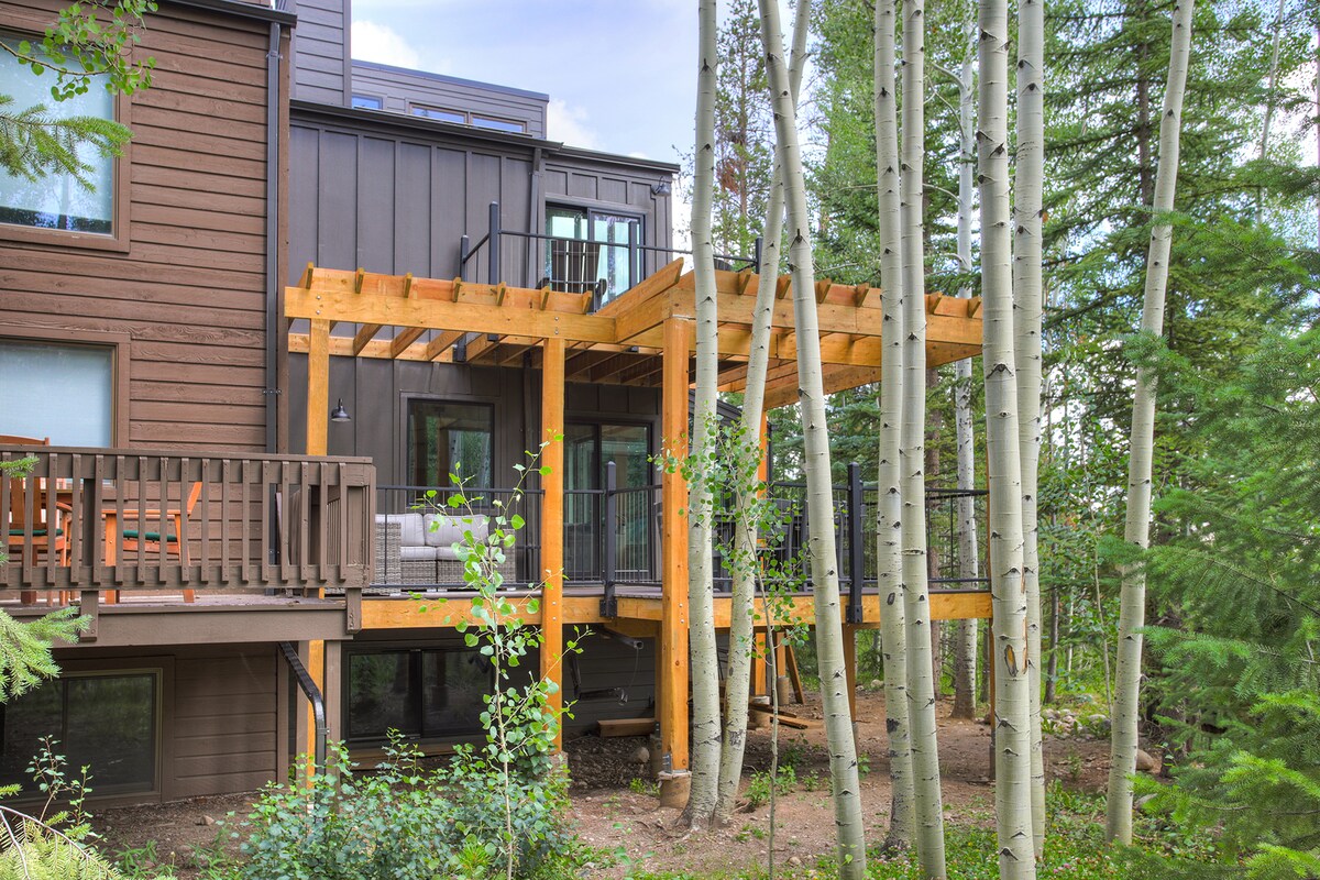 The outdoor deck features a wooden structure with ample seating and a shaded area, surrounded by tall aspen trees. The natural surroundings create a serene setting for relaxation, while large windows offer glimpses of the living space within.