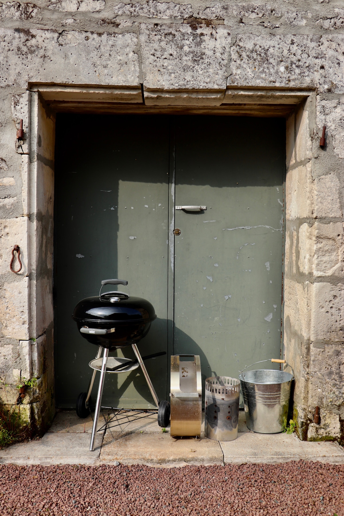 A black charcoal grill is positioned in front of a green door that features a simple metallic handle. Two metal containers are placed beside the grill, one cylindrical and the other rectangular, both suggesting outdoor cooking utility. The stone wall displays a rustic texture.