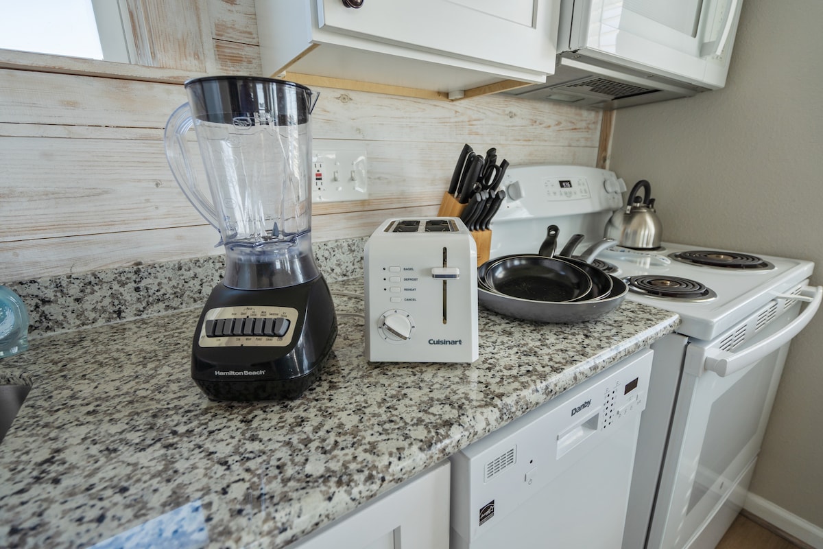 A well-equipped kitchen countertop displays a blender and a toaster, alongside frying pans and cooking utensils. The white appliances, including a dishwasher, are neatly arranged, complemented by the light-colored, textured wall behind the countertop.