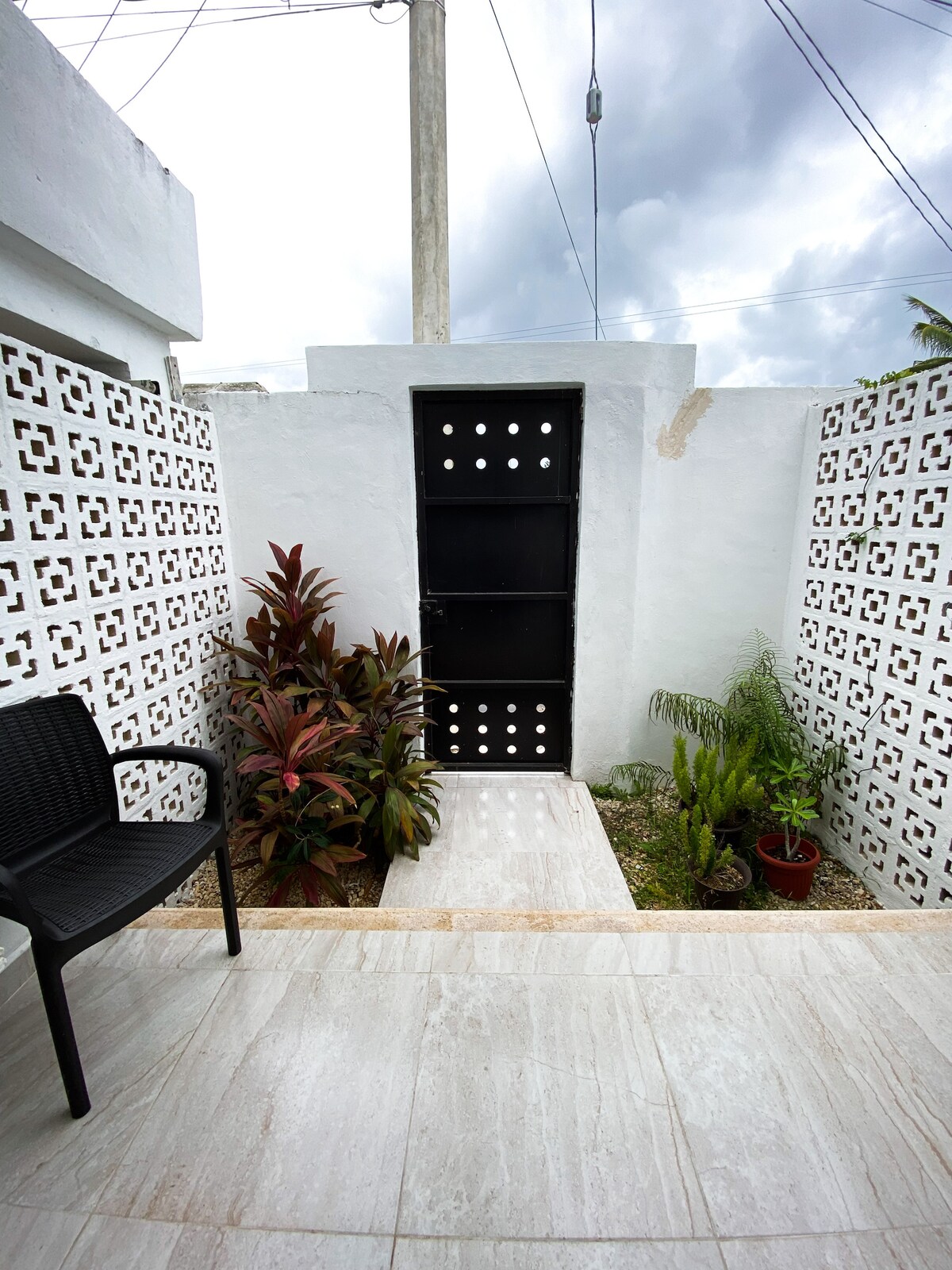 A simple outdoor area is presented with a black chair positioned next to a stone pathway. Lush green plants are arranged on both sides, complemented by a white wall with an intricate pattern. A solid black door leads into the space, framed by a cloudy sky.