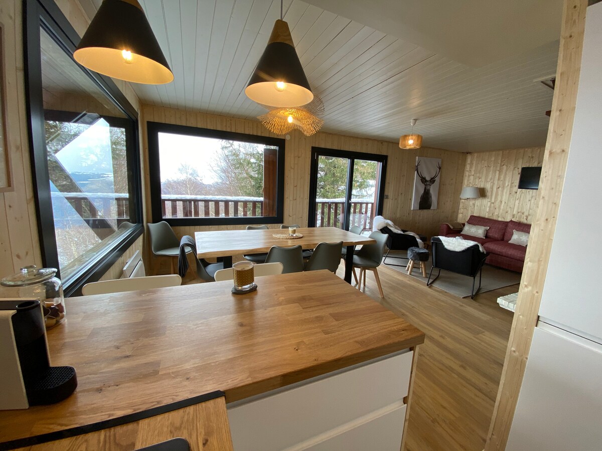 The open-plan living and dining area features a wooden table surrounded by six chairs. Large windows provide a panoramic view of the valley, while pendant lights hang above the table. A cozy seating area is arranged with a sofa and armchair in a warm color palette.