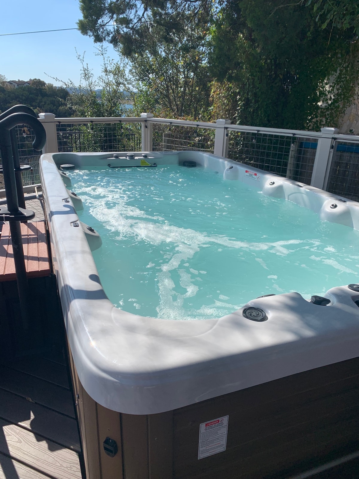 A private hot tub is situated on a deck, filled with clear blue water and equipped with jets. Surrounding foliage and a secure railing provide a sense of tranquility and seclusion, enhancing the outdoor relaxation experience.