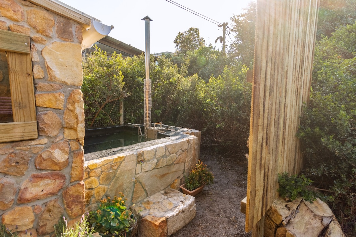 An outdoor stone wood-fired hot tub sits amid lush greenery, surrounded by rustic wooden features. Natural light filters through, highlighting the inviting water. Stone steps lead to the tub, enhancing the serene atmosphere of this secluded outdoor space.