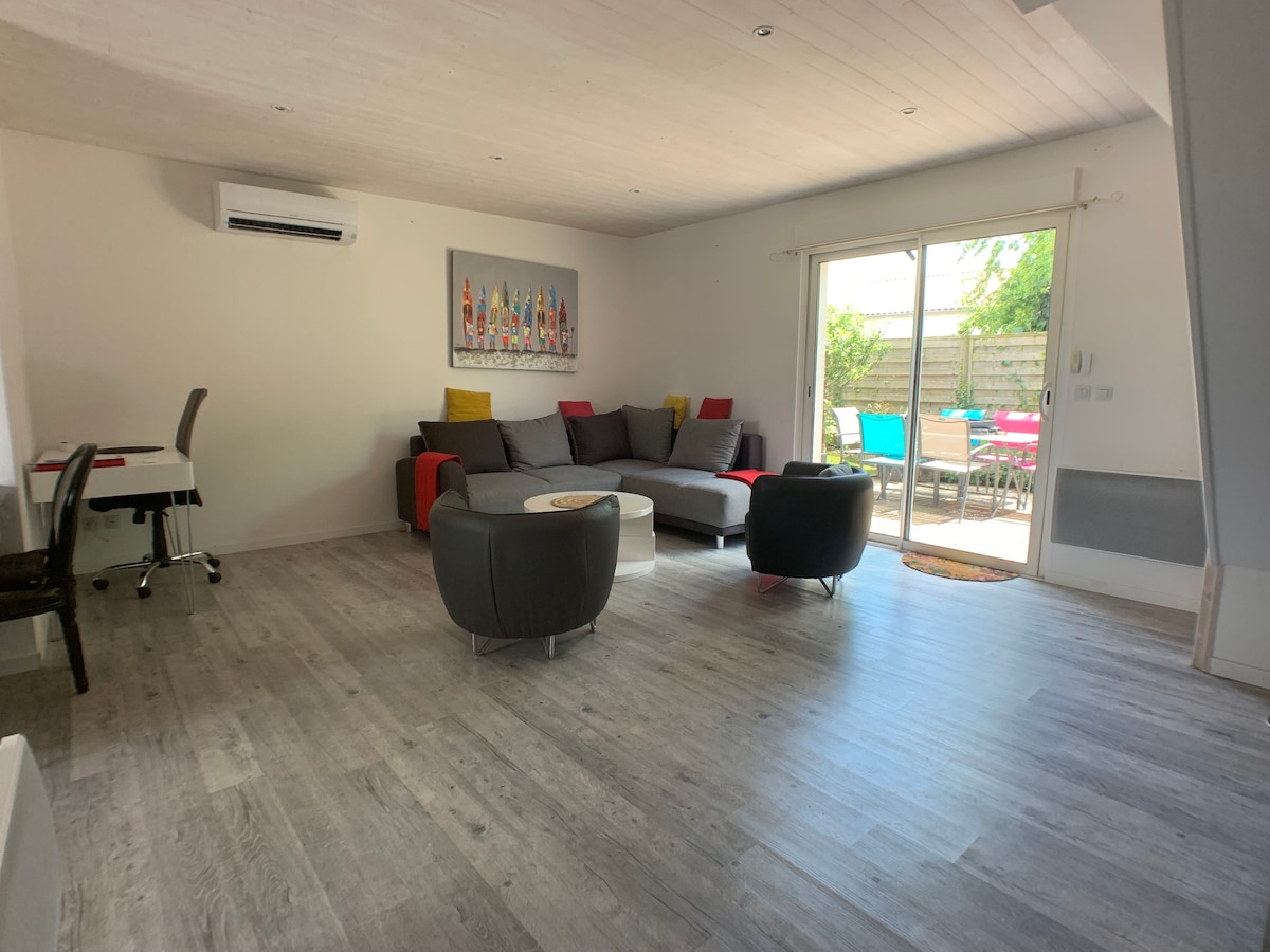 A bright living area features an air conditioning unit and a spacious corner sofa, accompanied by two comfortable armchairs. A dining table and chair are positioned nearby. Large glass doors open to a terrace, providing natural light and a view of outdoor seating.