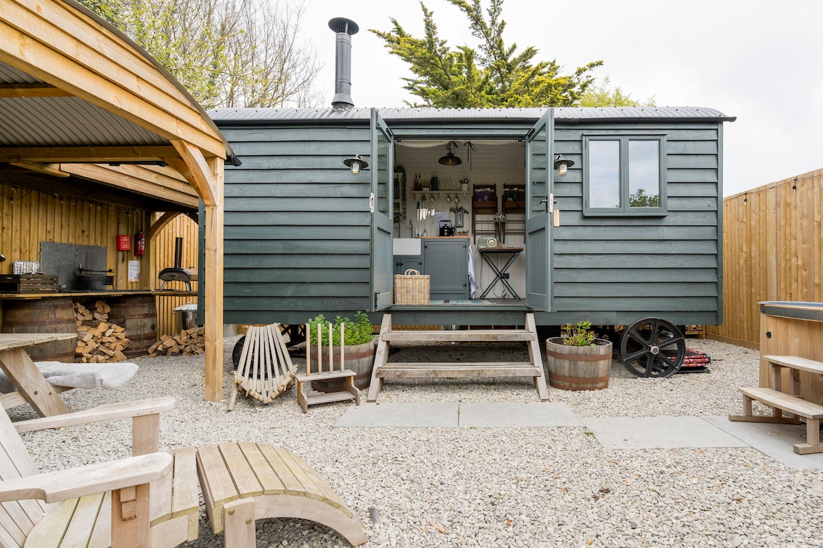 A charming hut features wide opening doors that invite light into the interior space. The surrounding area includes comfortable seating and a covered outdoor dining space, with a traditional wood-burning stove visible through the entrance. Gravel paths and planters add to the rustic landscape.