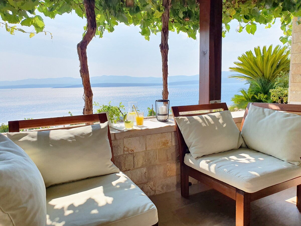 A shaded seating area is showcased with wooden chairs featuring soft cushions. A smooth stone wall is visible, adorned with lush greenery and a panoramic view of the tranquil sea. A lantern and two glasses sit on the table, enhancing the serene atmosphere.