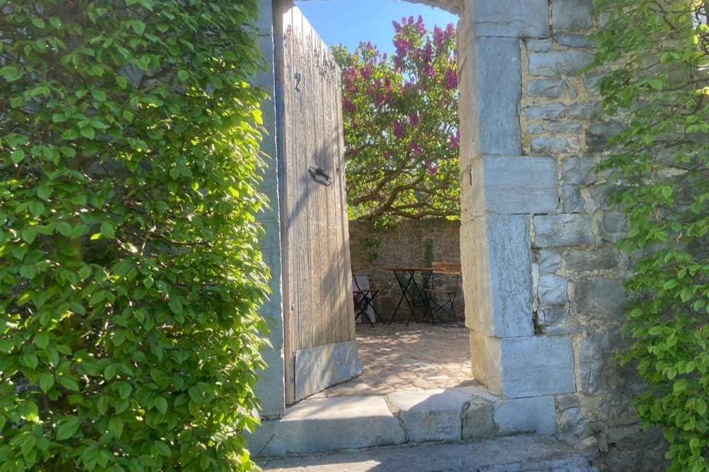 An open wooden gate frames a view of a quaint outdoor seating area, featuring a small round table and two chairs nestled among lush greenery. Blossoming trees in the background add a touch of color to the serene garden setting.